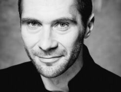 Czarno-biała fotografia. Uśmiechnięty mężczyzna z zarostem wpatruje się w obiektyw. Ubrany jest na czarno. Na zdjęciu: Piotr B. Dąbrowski.