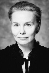Czarno-biała fotografia. Kobieta ze związanymi włosami wpatruje się w obiektyw. Ma na sobie bluzkę z koronkową kołnierzem. Na zdjęciu: Ewa Szumska.