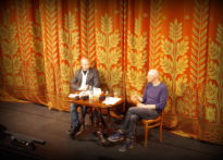 DWÓCH MĘŻCZYZN PRZY STOLIKU NA TLE OPUSZCZONEJ KURTYNY NA DUŻEJ SCENIE TEATRU POLSKIEGO W POZNANIU. OD LEWEJ PROFESOR PRZEMYSŁAW CZAPLIŃSKI I JAN SOWA. ZDJĘCIE ILUSTRACYJNE DO CYKLU NIE-BOSKIE NARRACJE. O LĘKU I GNIEWIE ROZMOWY Z PISARZAMI