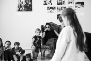 GRUPA MŁODZIEŻY SIEDZĄCEJ I STOJĄCEJ WOKÓŁ KOBIETY W CIEMNYCH OKULARACH NA TRONIE. PROJEKT LATO W TEATRZE "MIĘDZY SŁOWAMI" 2017