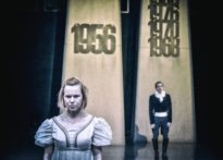 Na pierwszym planie aktorka w sukni, za nią aktor w białej koszuli z żabotem oraz surducie na tle repliki Poznański Krzyży