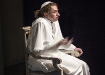 Aktor siedzący na krześle ubrany w strój papieski z gołymi stopami.