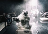 Na pierwszym planie postać przebrana za polarnego misia, na drugim publiczność oraz aktorzy.