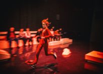 Kobieta biegnąca przez scenę, w oddali rozmazane postacie widzów