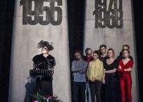 aktorka ubrana na czarno z woalką na twarzy stojąca przy pulpicie do nut, za nią pozostali aktorzy na tle Poznańskich Krzyży