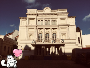 OŚWIETLONY SŁOŃCEM BUDYNEK TEATRU POLSKIEGO W POZNANIU. W LEWYM DOLNYM ROGU NAMALOWANA POSTAĆ GRUBEGO KOTA TRZYMAJĄCEGO W ŁAPKACH RÓŻOWY BALONIK W KSZTAŁCIE SERCA