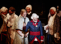 Siedmiu mężczyzn stojących na scenie w strojach epokowych i śmiejących się