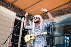 Aktor z peruką na głowie i ukulele przewieszonym przez szyję stojący na scenie z rękoma w górze