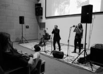 TRZECH MĘŻCZYZN NA SCENIE - DWÓCH PRZY MIKROFONACH, JEDEN GRA NA GITARZE. CZARNO BIAŁE ZDJĘCIE Z KONCERTU TRIO BRZOSKA MARCINIAK MARKIEWICZ