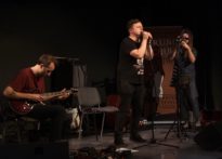 TRZECH MĘŻCZYZN NA SCENIE - DWÓCH PRZY MIKROFONACH, JEDEN GRA NA GITARZE. ZDJĘCIE ILUSTRACYJNE KONCERTU TRIO BRZOSKA MARCINIAK MARKIEWICZ