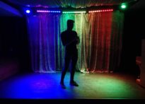 Stojący aktor z telefonem w ręce na tle tęczowej kotary. Na zdjęciu Piotr Dąbrowski