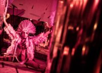 Aktor stojący w szpagacie na tle luster, ubrany w bieliznę, pierzastą marynarkę oraz indiański pióropusz na głowie. Na zdjęciu Krzysztof Broda- Żurawski