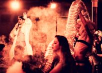 Aktorzy występujący wśród publiczności, mężczyzna z mikrofonem w centrum fotografii, pozostali aktorzy znajdują sie z boku niosąc element scenografii w kształcie rury.