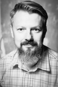 Czarno-biała fotografia. Mężczyzna w koszuli w kratę z wąsem i długą brodą. Na zdjęciu: dyrektor Marcin Kowalski.