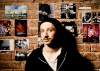Mężczyzna w czarnej czapce z wąsem opiera się o ceglaną ścianę, na której wiszą zdjęcia. Na zdjęciu Konrad Cichoń.